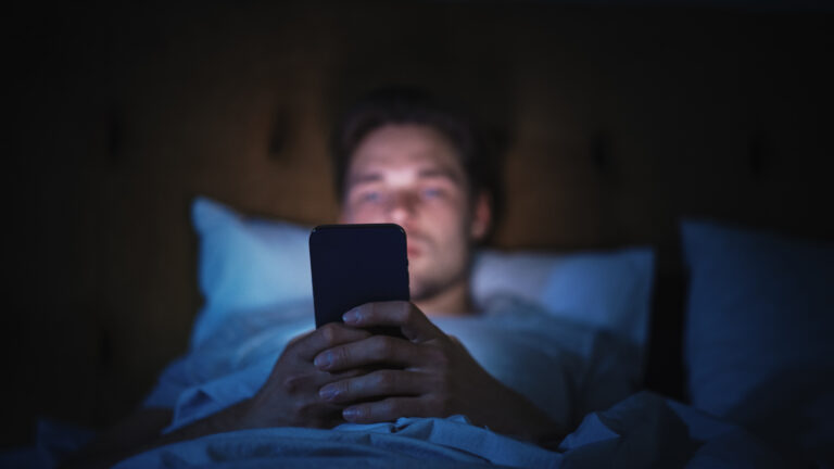 Caucasian Man Uses Smartphone in Bed at Home at Night. Handsome Guy Browsing Social Media, Reading News, Doing Online Shopping, Chatting with Friends Late at Night